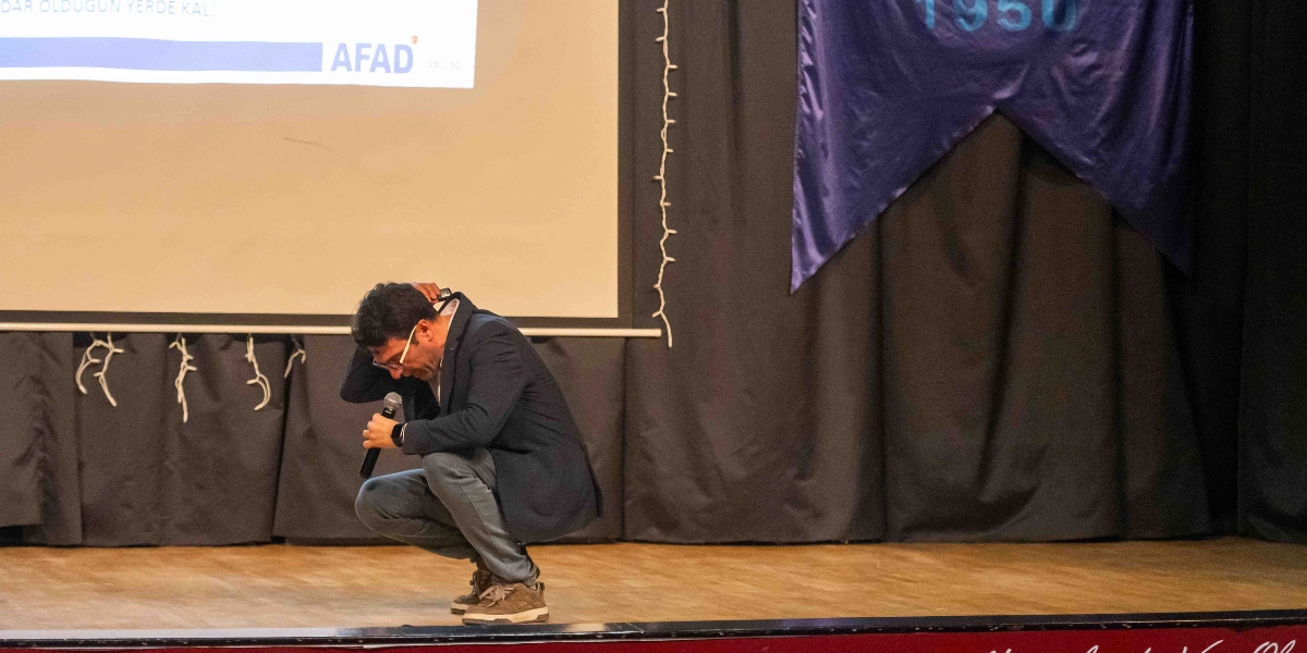 Bir konferans sahnesinde, lacivert ceketli ve gözlüklü bir eğitmen, elinde mikrofonla deprem anında yapılması gereken 