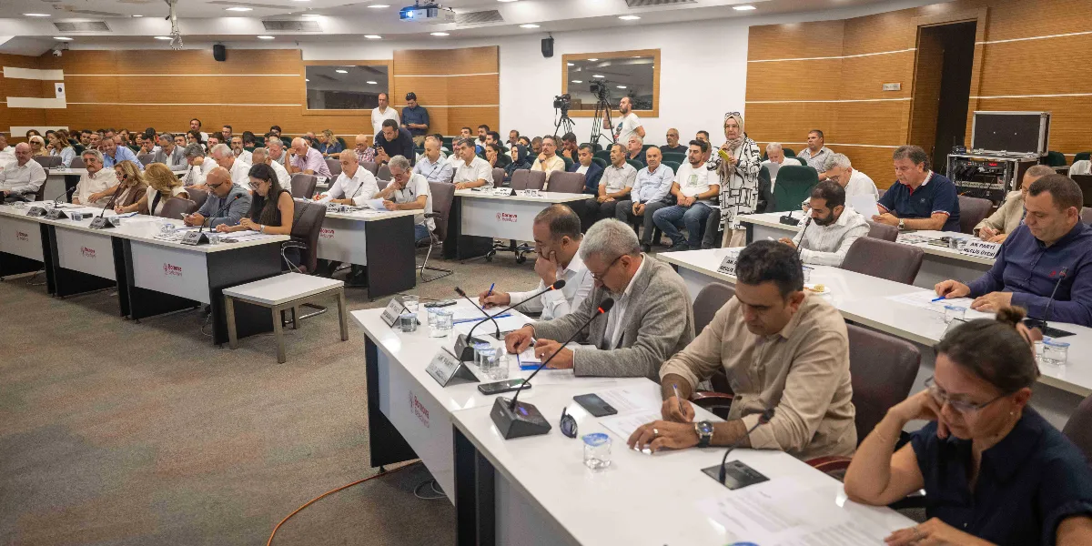 Bornova Belediyesi meclis salonunda düzenlenen toplantıda, sıralarda oturan çok sayıda katılımcı dikkatle not alıyor ve belgeleri inceliyor. Salonun ön planında masalarda mikrofonlar, isimlikler ve evraklar bulunurken, katılımcılar arasında Bornova Belediyesi logolu masalarda oturan meclis üyeleri yer alıyor. Arka planda ise ayakta duran veya oturan diğer katılımcılar ve teknik ekipmanlar görülüyor.