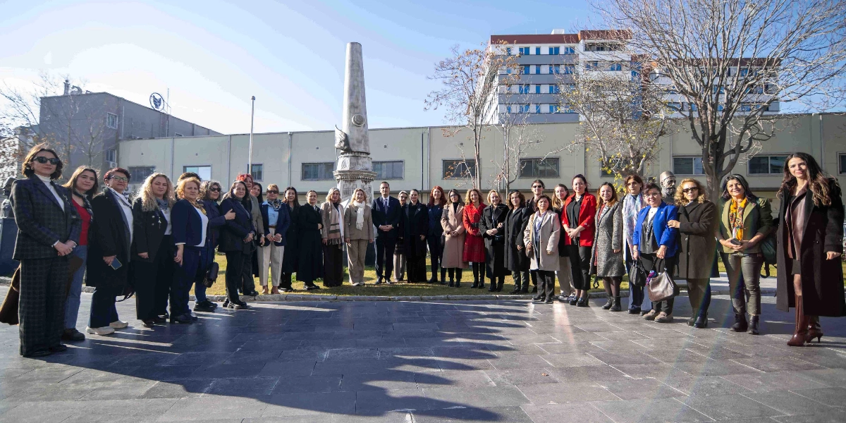 Açık havada, taş döşeli bir meydanda, arkalarında uzun bir anıt ve binaların bulunduğu alanda yan yana duran kalabalık bir grup kadın ve ortalarında yer alan bir erkekten oluşan topluluk. Gruptakiler günlük ve resmi kıyafetler içinde, kameraya gülümseyerek poz veriyor.