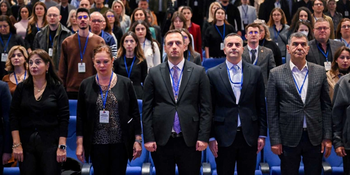 Resmi bir etkinlikte, boyunlarında etkinlik kartları asılı olan ve ciddiyetle karşıya bakan kalabalık bir grup insan. Ön sırada, takım elbiseli erkekler ve şık kıyafetli kadınlar yan yana durarak saygı duruşunda bulunuyor veya bir konuşmayı takip ediyor. Arka planda ise farklı yaş ve giyim tarzlarından oluşan, etkinliğe katılan çok sayıda kişi yer alıyor.