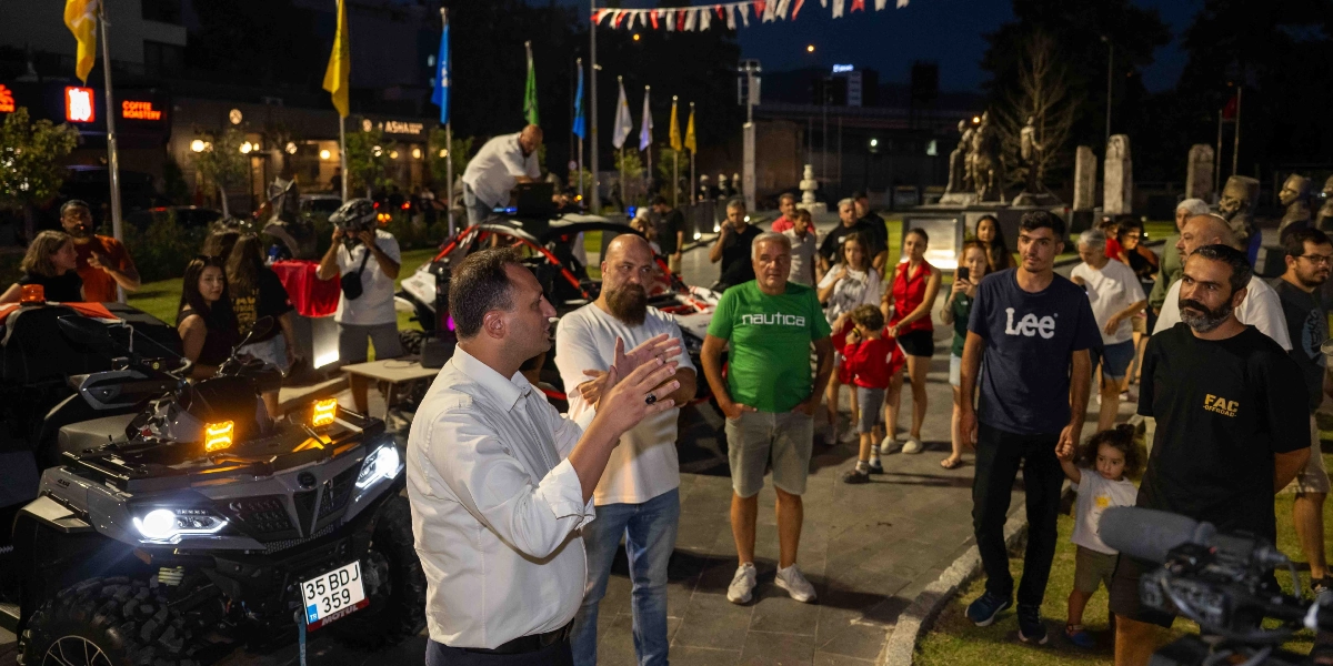 Akşam saatlerinde açık havada düzenlenen bir etkinlikte, beyaz gömlekli bir adam kalabalığa hitap ederken, yanında sakallı bir adam ve ön planda farları yanık, 35 BDJ 359 plakalı gri bir ATV yer alıyor. Etrafında farklı yaş gruplarından insanların toplandığı meydanda, arka planda bayraklar ve heykeller bulunuyor.