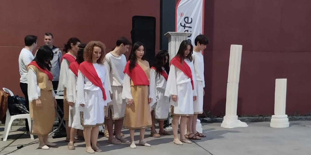 Antik Yunan dönemini anımsatan beyaz ve bej renkli tunikler giymiş, omuzlarında kırmızı şallar bulunan bir grup genç, başları hafifçe öne eğik şekilde yan yana duruyor. Arka planda bordo bir duvar, beyaz bir sütun parçası ve üzerinde 