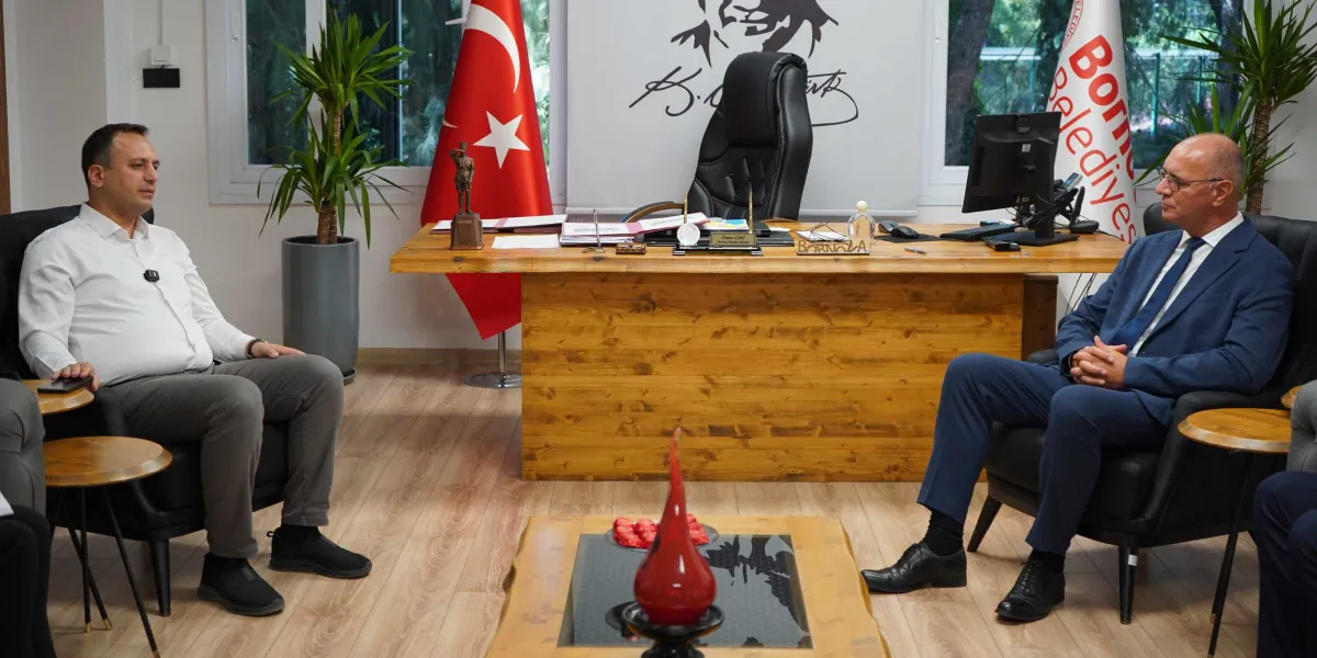 Bornova Belediye Başkanı Ömer Eşki, makam odasında Bosna-Hersek'ten gelen bir heyet temsilcisiyle karşılıklı oturarak görüşme gerçekleştiriyor. Arka planda Türk bayrağı, Atatürk imzalı bir tablo ve Bornova Belediyesi flaması yer alıyor. Ahşap çalışma masasının üzerinde küçük bir Atatürk heykeli ve isimlik bulunuyor.