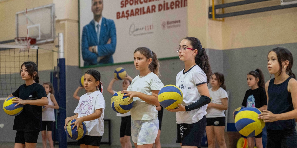 Kapalı bir spor salonunda voleybol antrenmanı yapan bir grup kız çocuğu, ellerinde voleybol toplarıyla yan yana dizilmiş şekilde duruyor. Çocuklar odaklanmış bir ifadeyle karşıya bakarken, arka planda Bornova Belediyesi'ne ait, üzerinde 
