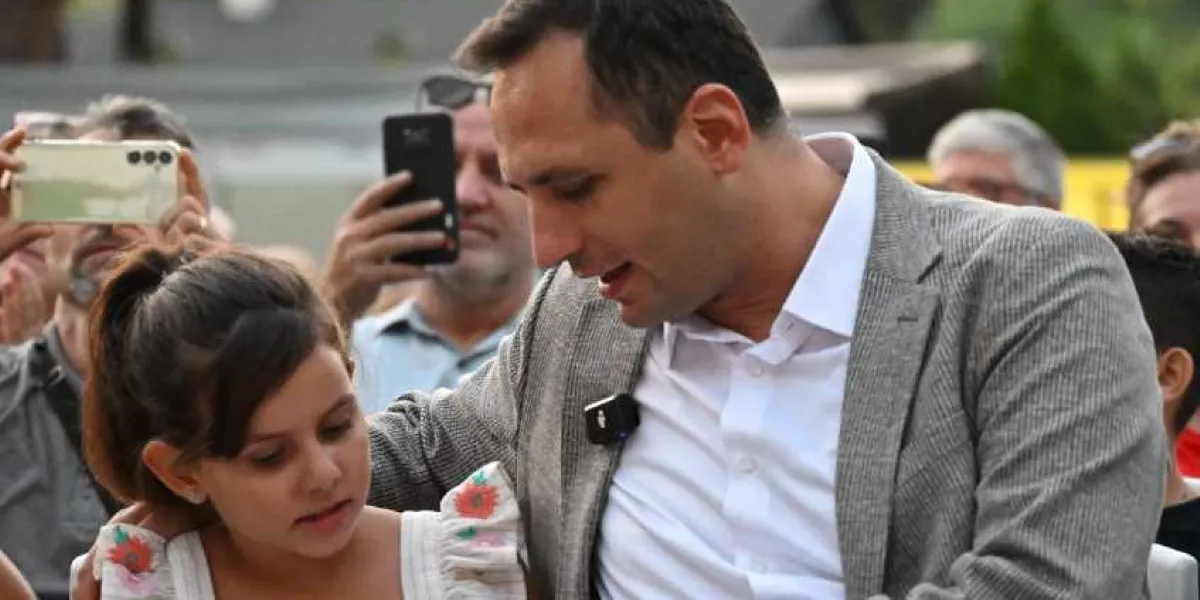 Gri ceketli ve beyaz gömlekli bir adam, yanında oturan küçük bir kız çocuğuna şefkatle bakarak onunla ilgileniyor. Arka planda ise etkinliği takip eden ve cep telefonlarıyla fotoğraf çeken bir kalabalık yer alıyor.