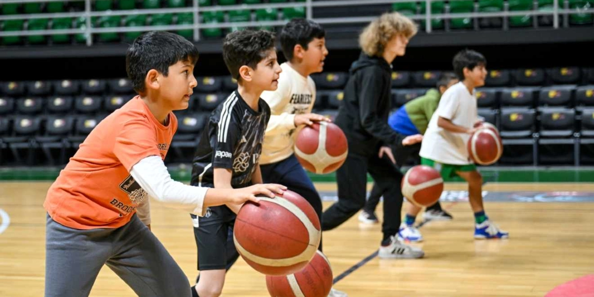 Kapalı bir spor salonunda basketbol antrenmanı yapan bir grup çocuk, ellerinde basketbol toplarıyla yan yana dizilmiş şekilde duruyor. Çocuklar, basketbol duruşu almış, odaklanmış bir şekilde ileriye bakıyorlar. Arka planda ise boş tribün koltukları yer alıyor.