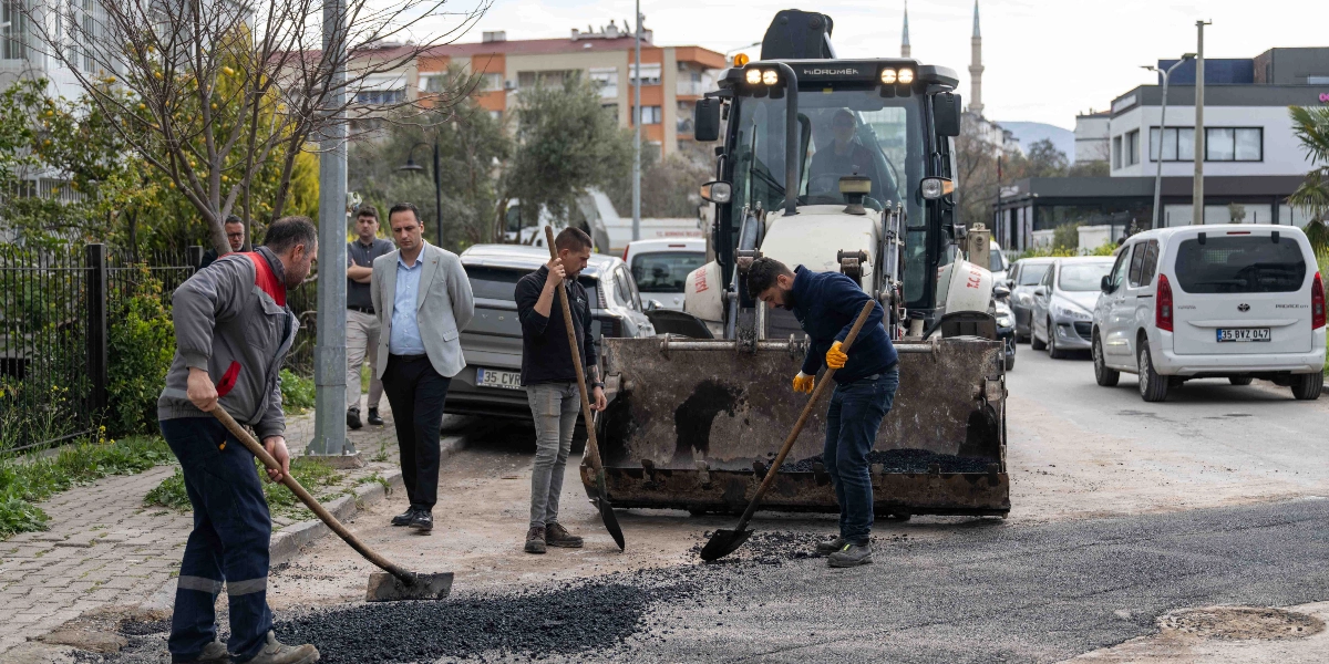 Bornova'da bir sokakta belediye ekipleri tarafından yürütülen yol yenileme çalışması. İşçiler ellerindeki küreklerle yola dökülen asfaltı düzeltirken, arkalarında büyük bir iş makinesi ve onu izleyen yetkililer bulunuyor. Arka planda park halindeki araçlar ve binalar yer alıyor.
