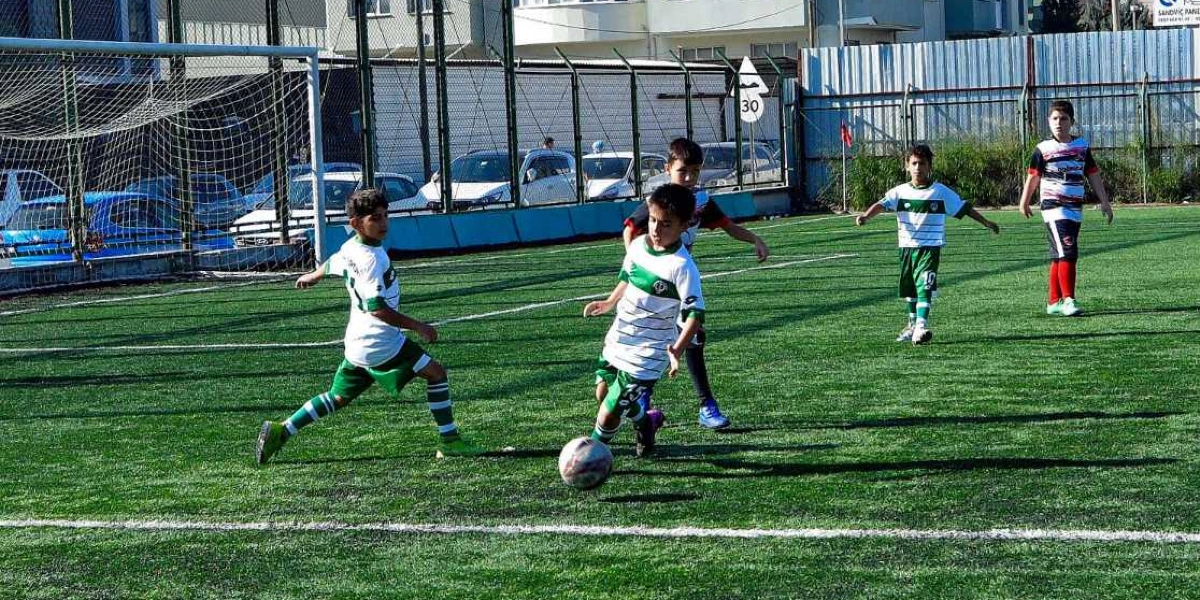 Yeşil çim sahada futbol oynayan çocuklardan biri topu sürerken, diğeri ona müdahale etmeye çalışıyor. Arka planda ise iki çocuk daha oyunu takip ediyor. Çocukların çoğu beyaz ve yeşil renkli formalar giyiyor, sağ tarafta ise farklı bir forma giyen bir çocuk daha bulunuyor. Sahanın çevresi tel örgülerle çevrili ve arka planda park halindeki araçlar görülüyor.