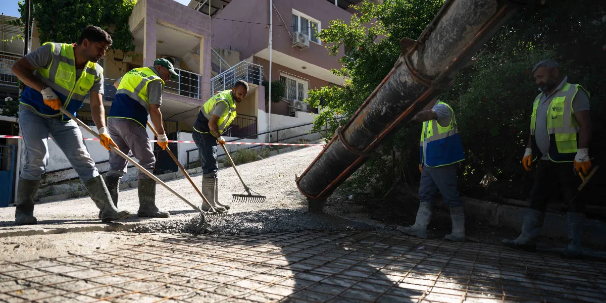 Bornova'da yokuşlu bir sokakta, üzerlerinde sarı reflektörlü yelekler ve çizmeler bulunan belediye işçileri, beton mikserinden dökülen harcı tırmık ve küreklerle yayarak yol çalışması yapıyor. İşçiler, metal hasır döşenmiş zemin üzerine beton dökme işlemini gerçekleştiriyor.