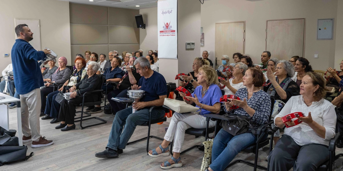 Bornova Belediyesi Altındağ Atatürk Kültür Merkezi'nde düzenlenen bir etkinlikte, eğitmen eşliğinde ellerinde tef ve darbuka gibi ritim aletleri bulunan katılımcılar müzik çalışması yapıyor. Salonun önünde duran eğitmen elindeki darbukayı çalarken, sandalyelerde oturan çoğunluğu orta yaş ve üzeri kadınlardan oluşan grup, ellerindeki kırmızı tefleri çalarak ona eşlik ediyor. Arka planda üzerinde 