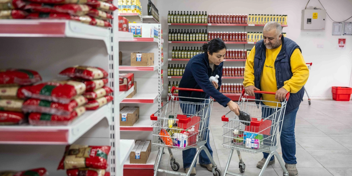 Bir market reyonunda alışveriş arabalarıyla bulunan bir kadın ve bir erkek, raflardan aldıkları gıda ürünlerini arabalarına yerleştiriyor. Raflarda düzenli bir şekilde dizilmiş yağ şişeleri, salça kutuları ve bakliyat paketleri görülüyor.