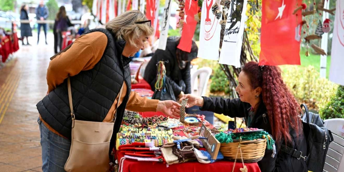 Açık havada kurulan bir hediyelik eşya pazarında, sarı kazak ve siyah yelek giyen sarışın bir kadın, tezgahın arkasında oturan kızıl saçlı satıcıdan el yapımı bir bileklik alırken görülüyor. Kırmızı örtüyle kaplı tezgahın üzerinde çeşitli takılar, nazar boncukları ve örgü ürünler sergileniyor. Arka planda ise üzerinde Atatürk silüeti ve Türk bayrakları bulunan beyaz flamalar asılı duruyor.
