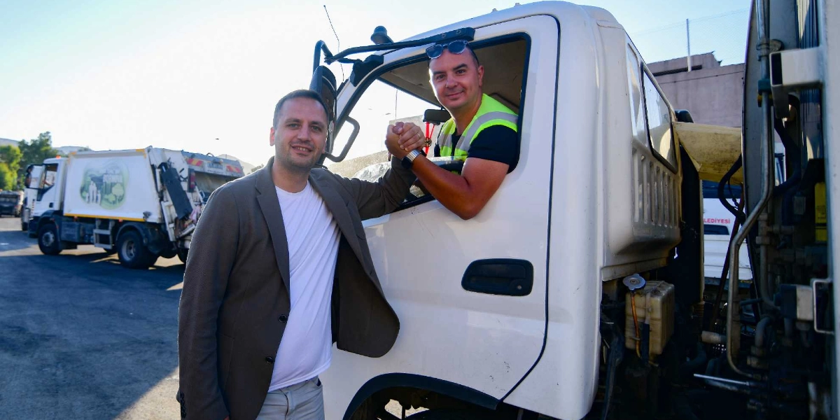 Beyaz tişört ve ceket giyen bir erkek, çöp kamyonunun şoför koltuğunda oturan ve üzerinde fosforlu iş yeleği bulunan bir başka erkekle el sıkışıyor. Arka planda Bornova Belediyesi logolu başka bir çöp kamyonu daha bulunuyor.