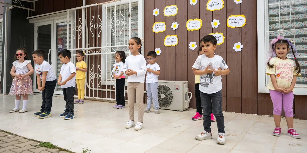 Bir anaokulunun bahçesinde yan yana dizilmiş, Anneler Günü için gösteri yapan neşeli çocuklar. Çocukların arkasındaki kahverengi duvarda, üzerinde 