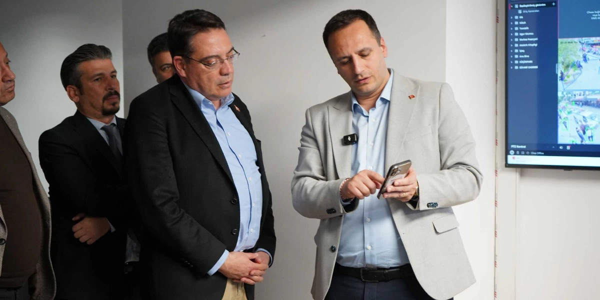 Açık renkli ceket giyen bir kişi, elindeki akıllı telefona bakarak yanındaki koyu renkli ceketli kişiye bir şeyler gösteriyor. İkili, duvarda asılı olan ve üzerinde güvenlik kamerası görüntüleri ile menü seçeneklerinin bulunduğu büyük bir ekranın önünde duruyor. Arka planda ise iki kişi daha onları izliyor.