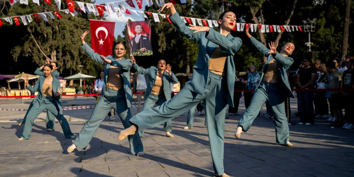 Açık havada, üzerinde Türk bayrakları ve Mustafa Kemal Atatürk posterinin asılı olduğu bir alanda, uyumlu mavi takım elbiseler ve bej renkli büstiyerler giymiş beş kadın dansçı modern bir koreografi sergiliyor. Dansçılar, kollarını ve bacaklarını estetik bir şekilde açarak dinamik bir figür oluşturuyor. Arka planda ise etkinliği izleyen bir grup seyirci bulunuyor.
