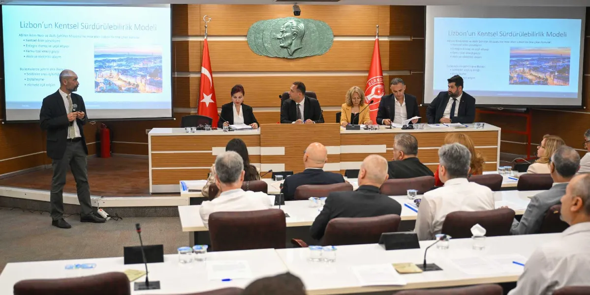Resmi bir toplantı salonunda, kürsüde sunum yapan bir konuşmacı ve onu dinleyen katılımcılar yer alıyor. Salonun ön tarafındaki masada beş kişilik bir heyet oturuyor; arkalarındaki duvarda ise Atatürk rölyefi ile Türk bayrağı ve Bornova Belediyesi'nin flaması bulunuyor. Yan taraflarda yer alan iki büyük projeksiyon ekranında 