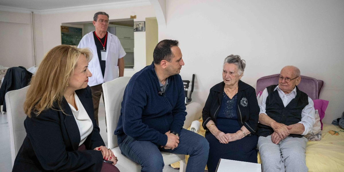 Aydınlık bir odada, orta yaşlı bir kadın ve bir erkek, yaşlı bir çiftle sohbet ediyor. Beyaz önlüklü bir erkek ise arka planda ayakta durarak onları izliyor. Yaşlı kadın ve erkek bir koltukta yan yana otururken, ziyaretçiler karşılarında sandalyelerde oturarak onlarla samimi bir şekilde konuşuyor.