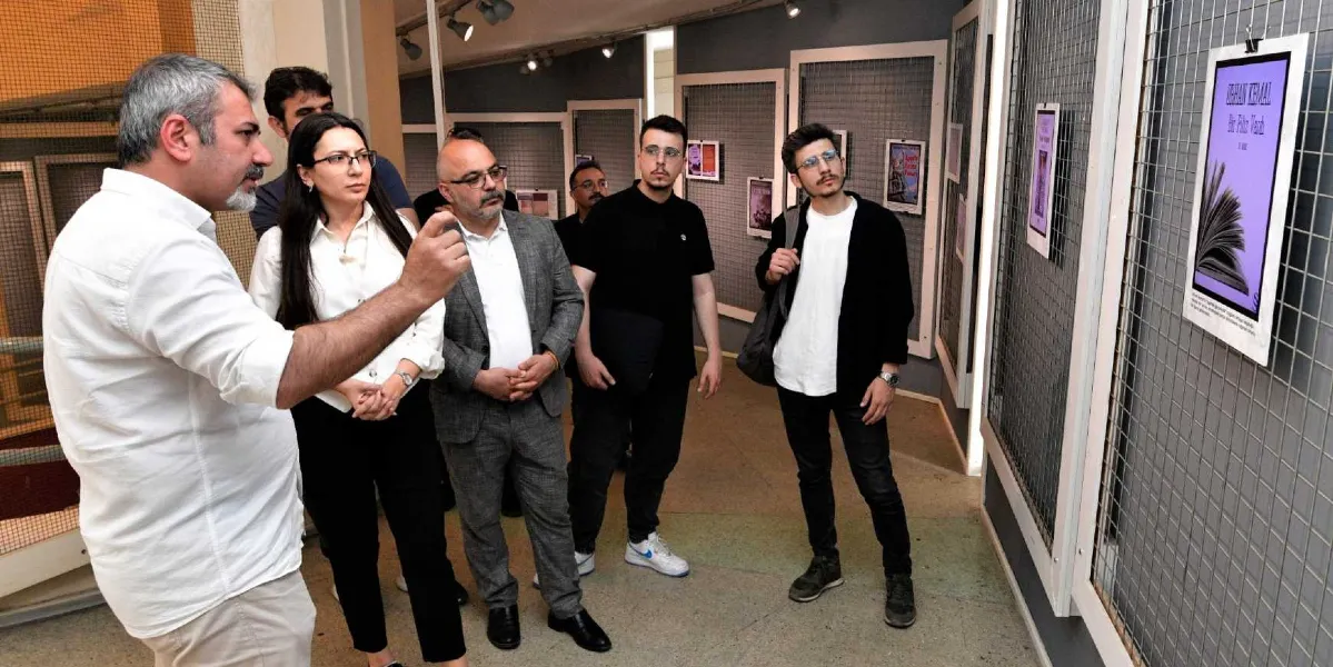 Bornova'da düzenlenen bir edebiyat sergisinde, bir grup ziyaretçi duvardaki tel panolara asılmış yazar ve kitap tanıtım afişlerini inceliyor. Ön planda beyaz gömlekli bir erkek, el hareketleriyle yanındaki gruba bilgi verirken, diğer katılımcılar dikkatle duvardaki 