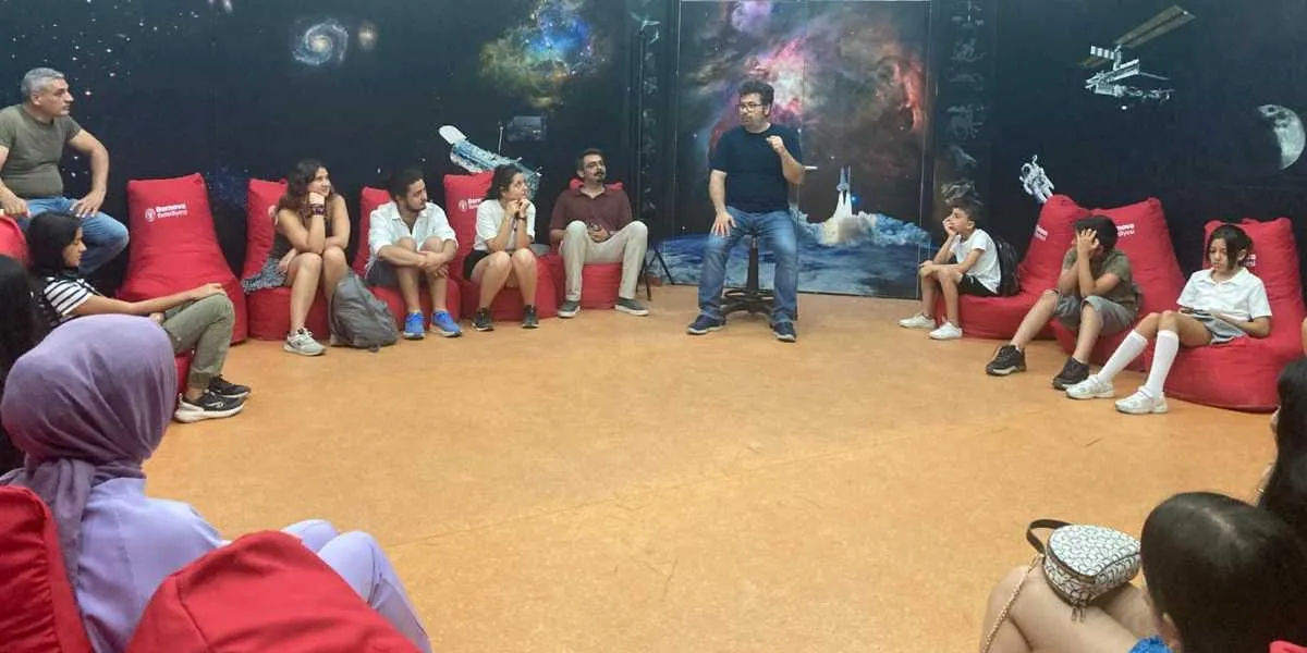 Uzay temalı duvar resimleriyle kaplı bir odada, kırmızı armut koltuklara oturmuş bir grup çocuk ve yetişkin, ortada bir taburede oturan konuşmacıyı dinliyor. Duvarlarda galaksiler, bir uzay istasyonu ve astronot figürleri yer alıyor.