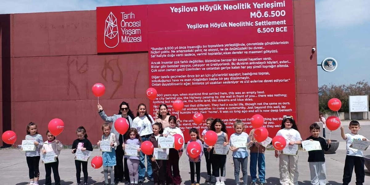 Yeşilova Höyük Neolitik Yerleşimi ve Tarih Öncesi Yaşam Müzesi yazılı büyük kırmızı bir duvarın önünde duran, ellerinde kırmızı balonlar ve katılım belgeleri tutan bir grup çocuk ve onlara eşlik eden yetişkinler. Duvarın üzerinde, bölgenin tarihini anlatan Türkçe ve İngilizce bilgilendirici metinler yer alıyor.