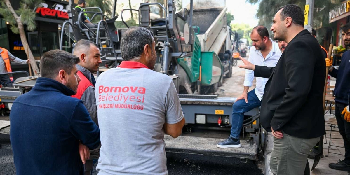 Bornova Belediyesi Fen İşleri Müdürlüğü personeli ve yetkililer, Süvari Caddesi'ndeki yol yenileme çalışmalarını yerinde inceliyor. Arka planda asfalt serme makinesi ve damperli kamyon bulunurken, ön planda sırtında belediye logosu olan bir görevli ve yanındaki diğer çalışanlar, bir yetkilinin çalışmaları anlattığı sırada onu dinliyor.