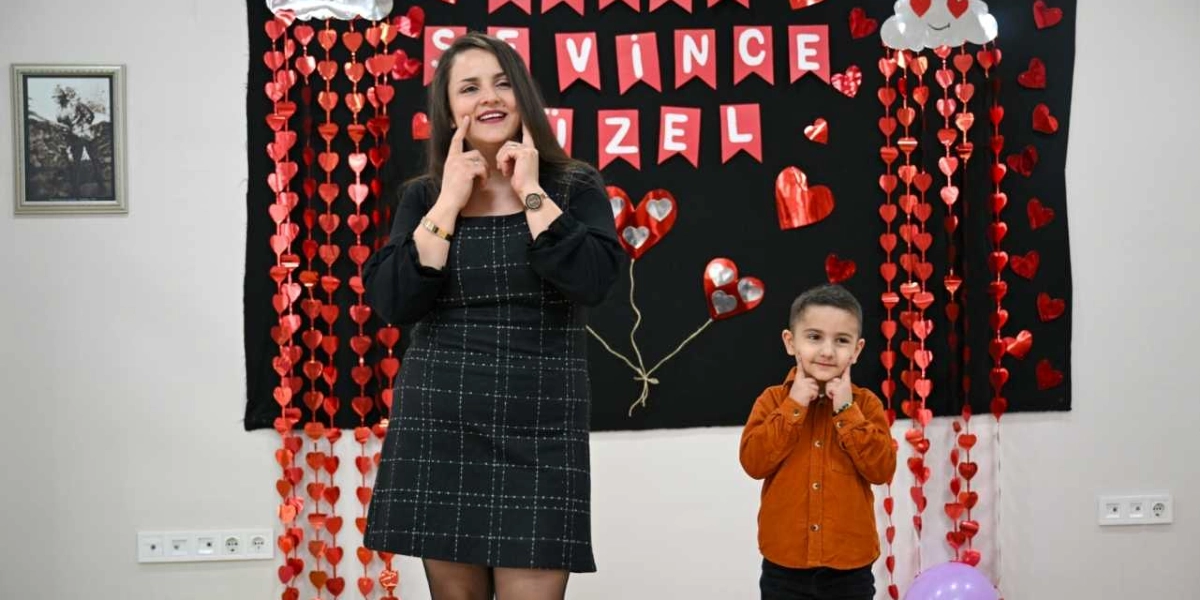Siyah kareli bir elbise giymiş bir kadın ve turuncu gömlekli küçük bir erkek çocuk, arkalarında kalp süslemeleri ve üzerinde 