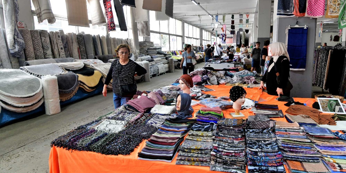 Kapalı bir pazar yerinde, turuncu örtülü uzun bir tezgahın üzerinde düzenli bir şekilde dizilmiş çok sayıda renkli eşarp ve şal sergileniyor. Tezgahın üzerinde ürünlerin sergilendiği iki adet cansız manken başı bulunuyor. Tezgahın çevresinde alışveriş yapan insanlar ve arka planda duvar boyunca rulo halinde dizilmiş halılar ile çeşitli tekstil ürünleri yer alıyor.