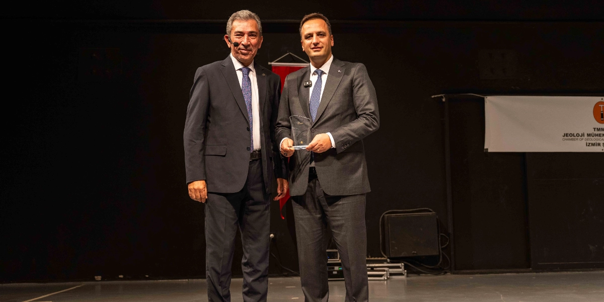 Koyu renkli bir sahnede, takım elbiseli iki erkek yan yana duruyor. Sağdaki kişi, elinde şeffaf bir plaket tutuyor. Arka planda TMMOB Jeoloji Mühendisleri Odası İzmir Şubesi'ne ait bir pankart yer alıyor.