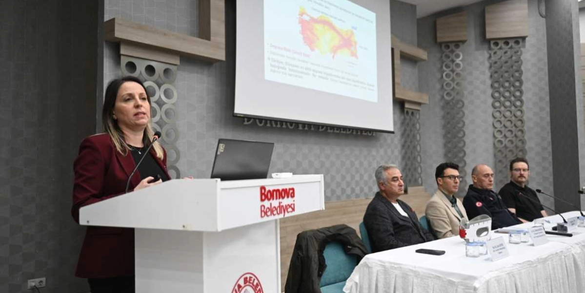 Bornova Belediyesi kürsüsünde konuşma yapan bir kadın ve onu dinleyen dört kişilik bir panelist grubu. Konuşmacının arkasındaki projeksiyon ekranında Türkiye'nin deprem risk haritası yer alıyor. Panelistler masada otururken, konuşmacı elinde bir sunum kumandasıyla izleyicilere hitap ediyor.