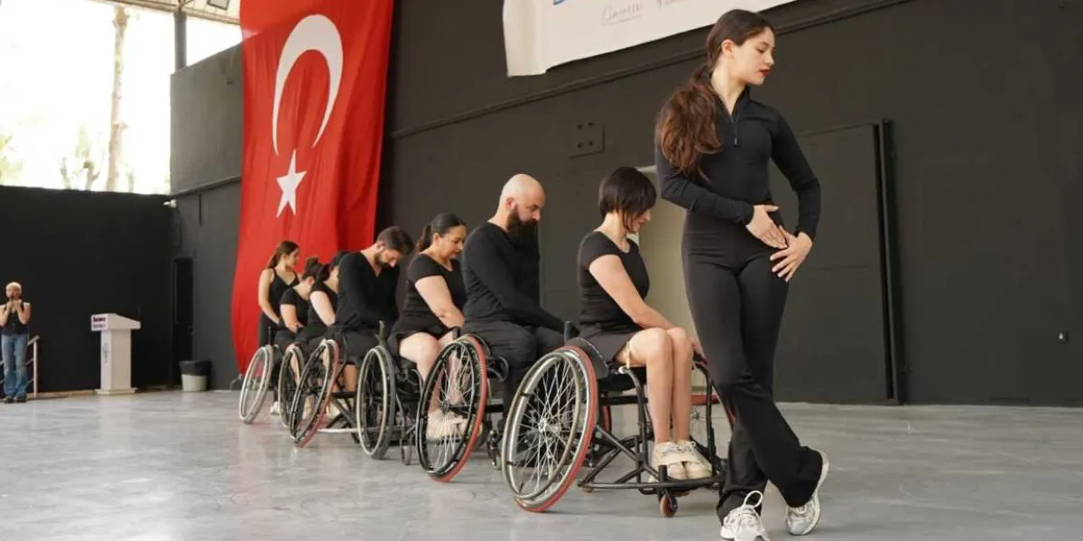 Siyah kıyafetler giymiş bir grup dansçı, kapalı bir spor salonunda yan yana dizilmiş tekerlekli sandalyelerde oturuyor. Grubun en önünde ayakta duran genç bir kadın dansçı, zarif bir duruşla onlara eşlik ediyor. Arka planda büyük bir Türk bayrağı asılı duruyor ve sol tarafta bir kürsü ile bir kişi daha görülüyor.