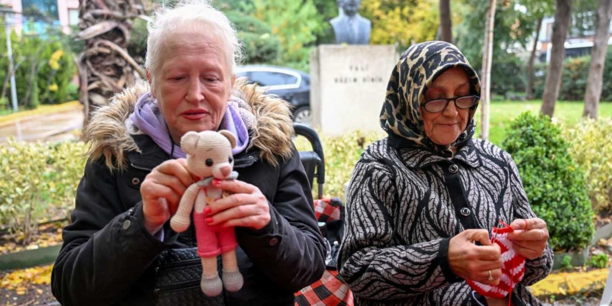 Açık havada, bir park ortamında yan yana oturan iki kadın el işi yapıyor. Soldaki kadın, kürklü kapüşonu olan siyah bir mont giymiş ve elinde örgüden yapılmış küçük bir oyuncak ayı tutuyor. Sağdaki kadın ise leopar desenli bir başörtüsü ve siyah beyaz desenli bir hırka giymiş, elinde kırmızı beyaz iplerle örgü örüyor. Arka planda ağaçlar, bir büst ve parkta park etmiş bir otomobil görülüyor.