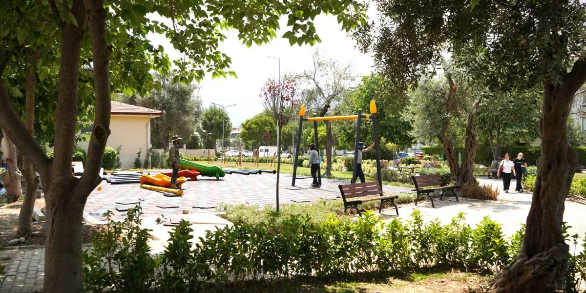 Ağaçlarla çevrili bir park alanında, zemin döşemesi devam eden oyun parkı kurulumu görülüyor. Parkın ortasında montajı süren renkli oyun grubu parçaları, spor aletleri, ahşap banklar ve yürüyüş yolları yer alıyor. Arka planda çalışan işçiler ve parkta yürüyen insanlar bulunuyor.