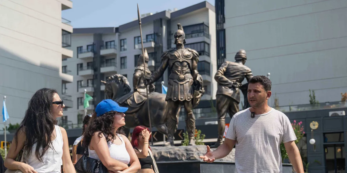 Abide-i Hürriyet Meydanı'nda, elinde mızrak tutan miğferli bir savaşçı ve atlı figürlerin yer aldığı bronz heykellerin önünde duran bir grup insan. Ön planda, çizgili tişörtlü bir erkek, el hareketleriyle bir şeyler anlatırken, yanında duran genç kadınlar heykellere bakıyor. Arka planda ise modern apartman binaları ve peyzaj düzenlemesi görülüyor.