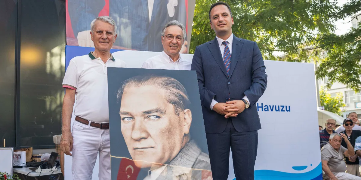 Bornova Belediye Başkanı Mustafa İduğ ve beraberindeki iki kişi, üzerinde Mustafa Kemal Atatürk'ün portresinin yer aldığı bir panoyu tutarak poz veriyor. Arka planda üzerinde 