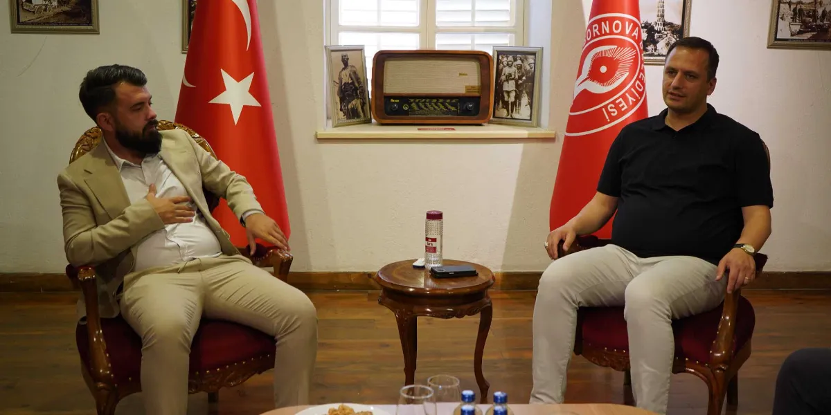 Bej renkli takım elbiseli, sakallı bir erkek ile siyah polo yaka tişört ve açık renkli pantolon giyen bir erkek, aralarında küçük bir sehpa bulunan bordo koltuklarda karşılıklı oturarak sohbet ediyor. Arka planda ortada eski bir radyo, iki yanında tarihi fotoğraflar, yanlarda ise Türk bayrağı ve üzerinde Bornova Belediyesi logosu bulunan kırmızı bir flama yer alıyor.