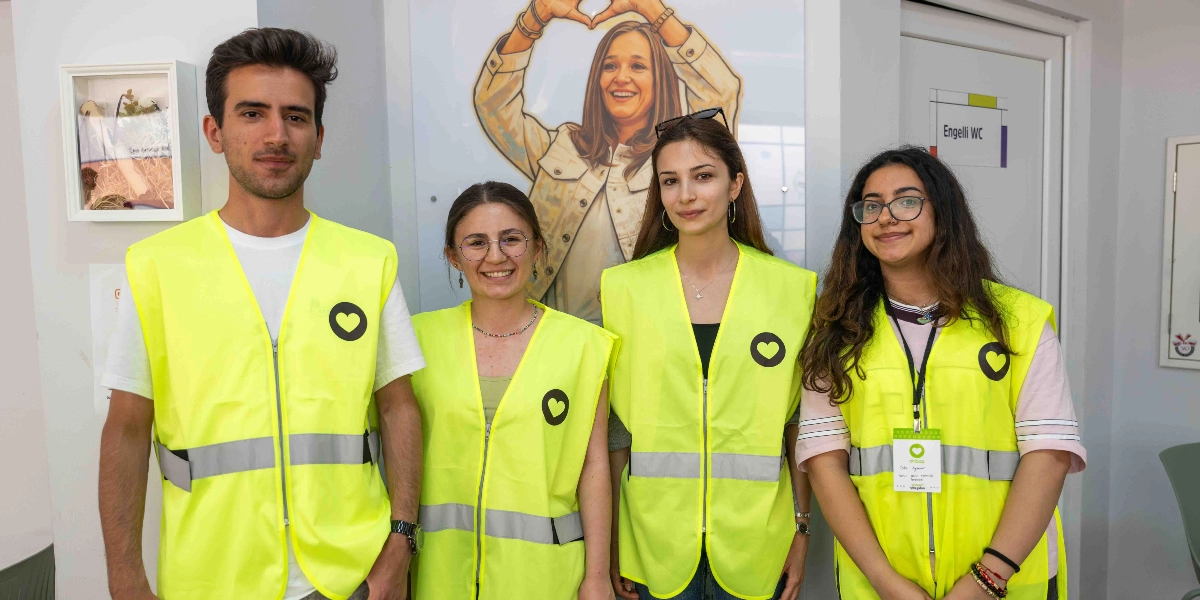 Üzerinde siyah kalp logosu bulunan fosforlu sarı yelekler giymiş iki erkek ve iki kadın yan yana durarak gülümsüyor. Arka planda elleriyle kalp işareti yapan bir kadın figürünün olduğu duvar görseli ve üzerinde 