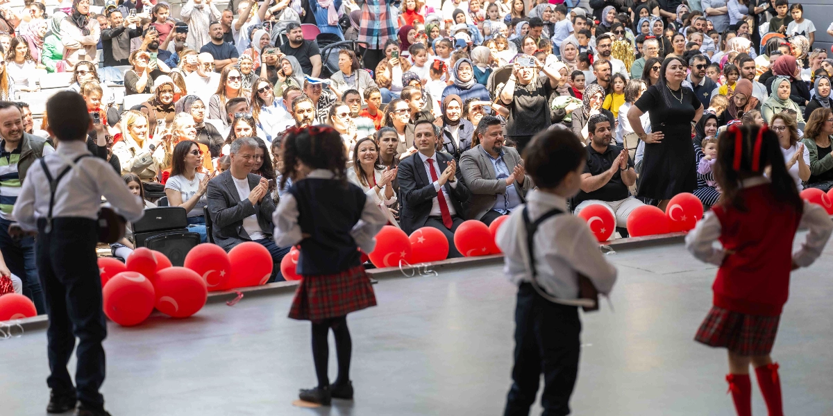 23 Nisan Ulusal Egemenlik ve Çocuk Bayramı kutlamaları kapsamında sahnede halk oyunları kıyafetleri giymiş dört çocuk dans ederken, arkalarındaki tribünlerde oturan kalabalık bir izleyici grubu onları alkışlıyor. Sahnenin ön kısmında kırmızı Türk bayraklı balonlar dizili duruyor, izleyiciler arasında ise resmi kıyafetli yetişkinler dikkat çekiyor.