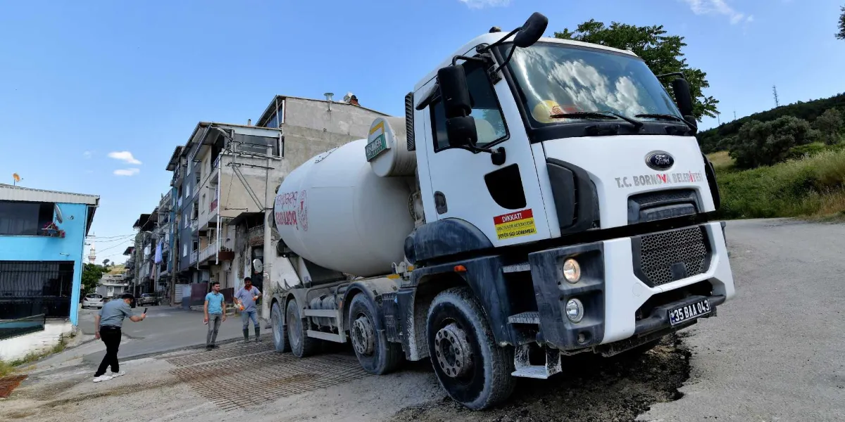 Üzerinde T.C. Bornova Belediyesi yazan beyaz bir beton mikseri, yerleşim yerindeki bir yol çalışmasında beton dökümü yapıyor. Kamyonun ön kısmında 
