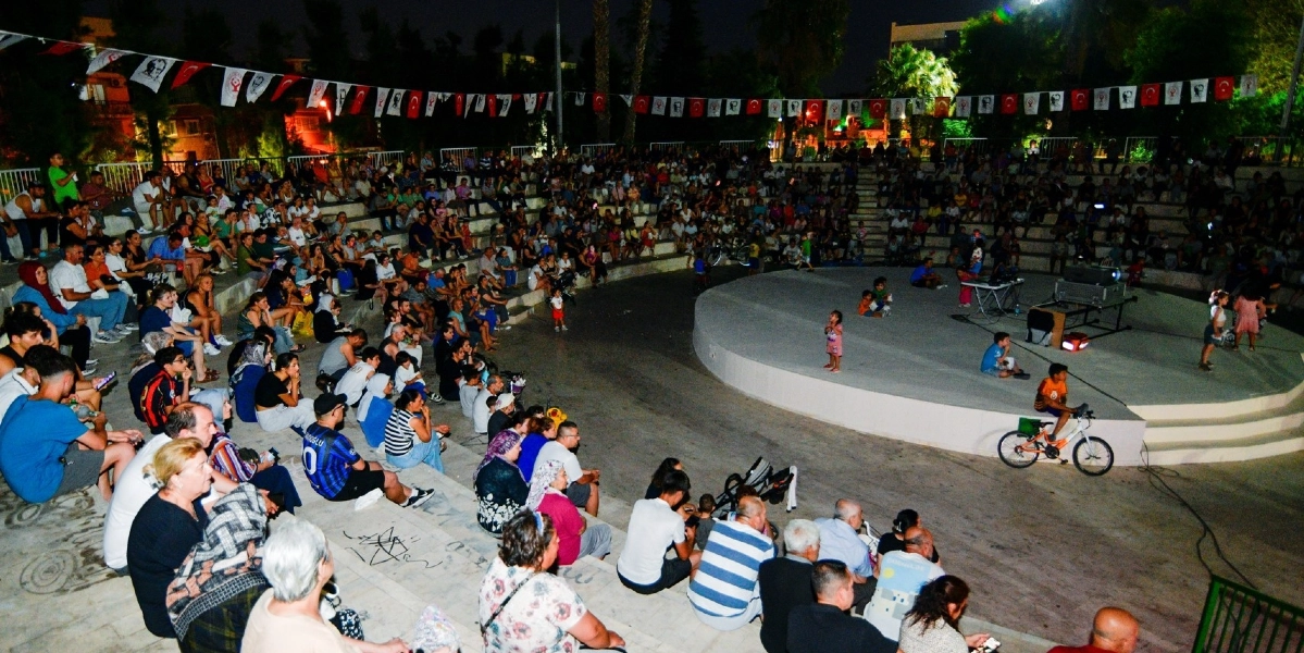 Gece vakti açık hava amfitiyatrosunda toplanmış kalabalık bir izleyici grubu, sahneye doğru dönük şekilde beton basamaklarda oturuyor. Sahnenin üzerinde bir projeksiyon cihazı ve teknik ekipmanlar bulunurken, bazı çocuklar sahne alanında oyun oynuyor. Alanın çevresi, üzerinde Türk bayrakları ve çeşitli logoların bulunduğu iplere asılı flamalarla süslenmiş.