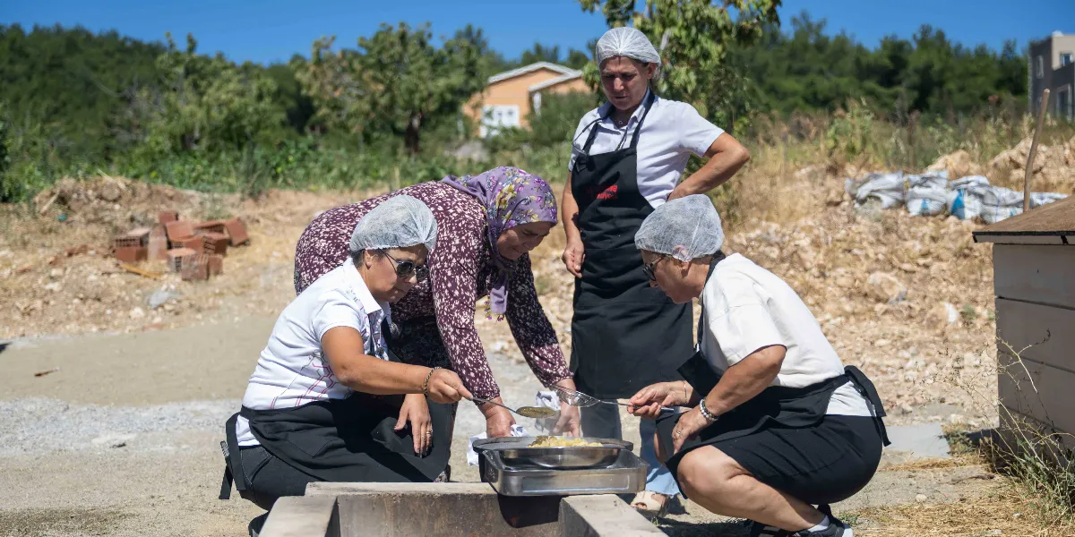 Açık havada, üzerinde beyaz hijyenik boneler ve siyah önlükler bulunan dört kadın, yere yerleştirilmiş bir ocak üzerinde yemek pişiriyor. Kadınlardan ikisi çömelmiş vaziyette ellerindeki süzgeçlerle tavadaki yiyecekleri karıştırırken, diğer ikisi onları izliyor. Arka planda ağaçlık bir alan ve müstakil bir ev görülüyor.