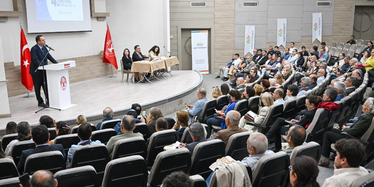 Modern bir konferans salonunda gerçekleşen Bornova Kent Konseyi Genel Kurul toplantısında, kürsüde konuşma yapan bir yetkili ve onu dinleyen kalabalık bir izleyici grubu yer alıyor. Sahnedeki kürsünün yanında Türk bayrağı ve Bornova Belediyesi bayrağı bulunurken, arka planda üzerinde 
