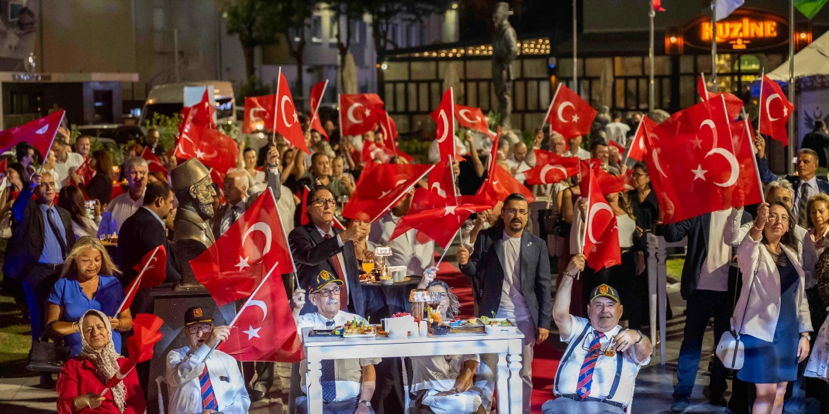 Gece vakti düzenlenen açık hava resepsiyonunda, ellerinde Türk bayrakları taşıyan kalabalık bir grup coşkuyla kutlama yapıyor. Ön planda, beyaz bir masanın etrafında oturan, üzerinde askeri şapkalar ve kravatlar bulunan iki yaşlı beyefendi ve yanlarında ayakta duran katılımcılar yer alıyor. Arka planda ise bir Atatürk büstü, ışıklandırılmış bir restoran binası ve resepsiyona katılan çok sayıda davetli görülüyor.
