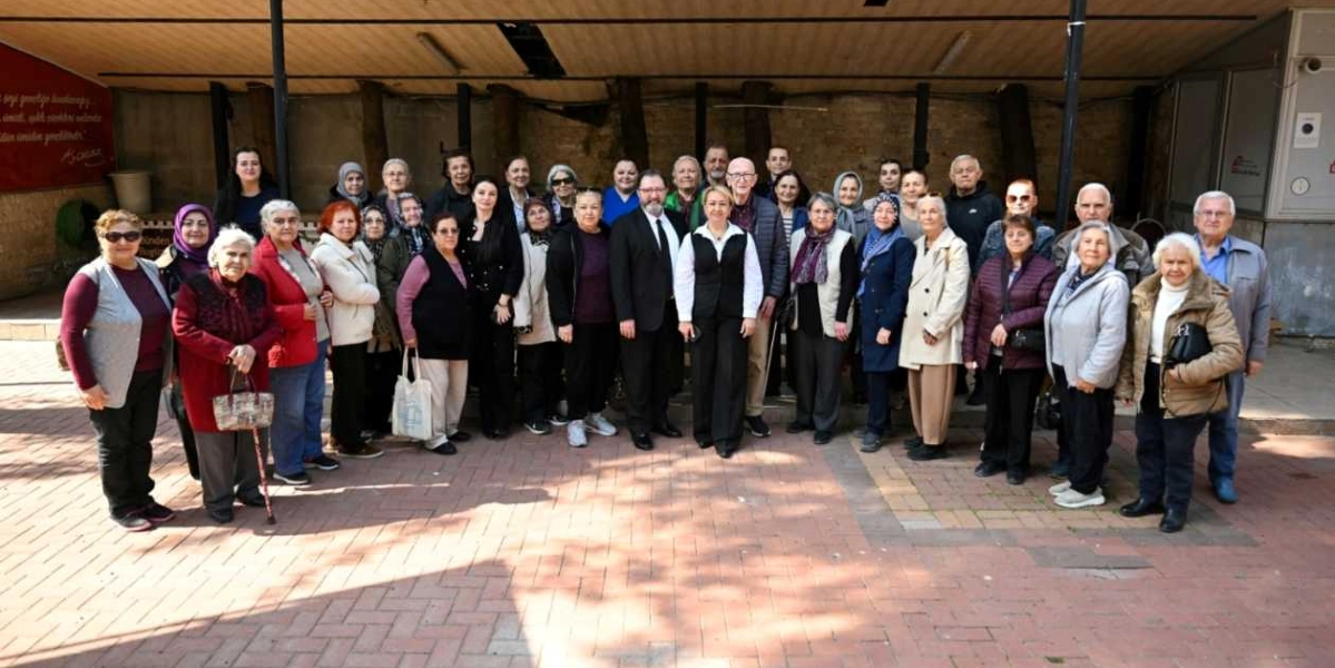 Bornova'da düzenlenen bir etkinlikte, çoğunluğu yaşlı bireylerden oluşan kalabalık bir grup, açık havada yan yana gelerek hatıra fotoğrafı çektiriyor. Grubun orta kısmında, siyah takım elbiseli bir erkek ve beyaz gömlek üzerine siyah yelek giymiş bir kadın yer alıyor. Katılımcılar günlük kıyafetleriyle, gülümseyerek objektife bakıyor. Arka planda ise taş duvarlı ve sundurmalı bir yapı bulunuyor.