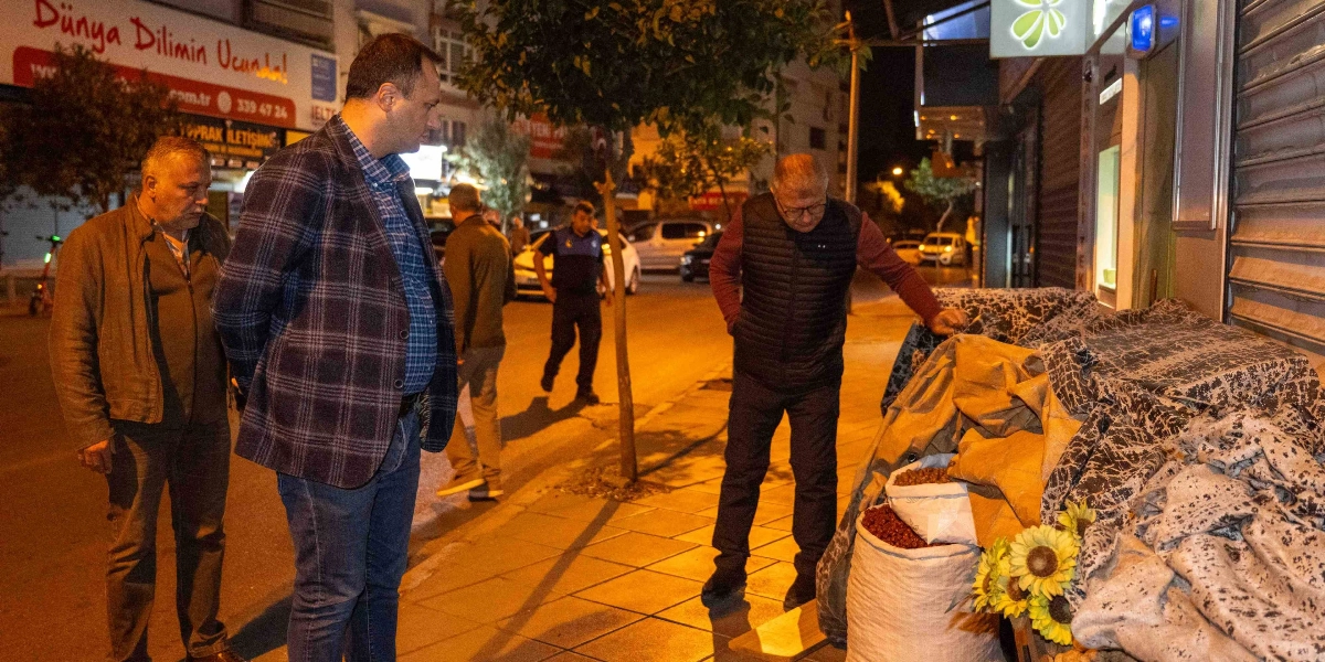 Gece vakti bir sokakta, kaldırımı işgal eden eşyaların başında duran Bornova Belediye Başkanı Mustafa İduğ ve beraberindeki yetkililer, kaldırımın yayalar için temizlenmesi sürecini denetliyor. Kaldırım üzerinde kumaşlarla örtülü tezgahlar, çuvallar ve yapay ayçiçekleri bulunuyor.