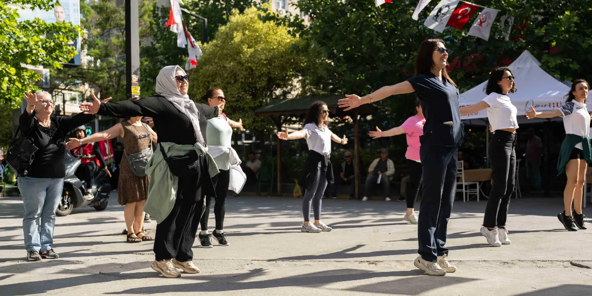 Açık havada, park gibi bir alanda bir araya gelmiş farklı yaş ve giyim tarzlarından oluşan bir grup kadın, kollarını iki yana açarak egzersiz yapıyor. Arka planda ağaçlar, Türk bayrakları ve üzerinde 