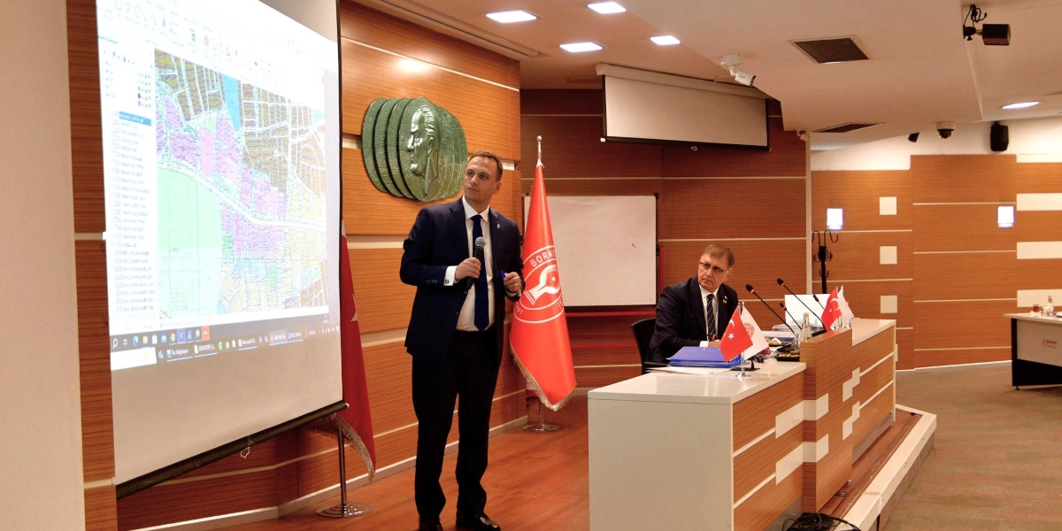 Modern bir toplantı salonunda, takım elbiseli bir konuşmacı elinde mikrofonla projeksiyon ekranındaki detaylı bir şehir planlama haritasını sunuyor. Konuşmacının arkasındaki duvarda Atatürk rölyefi ve Bornova Belediyesi'nin kırmızı renkli bayrağı yer alıyor. Masada oturan bir diğer yetkili ise sunumu dikkatle takip ediyor; masanın üzerinde Türk bayrağı ve belediye bayrağı bulunuyor.