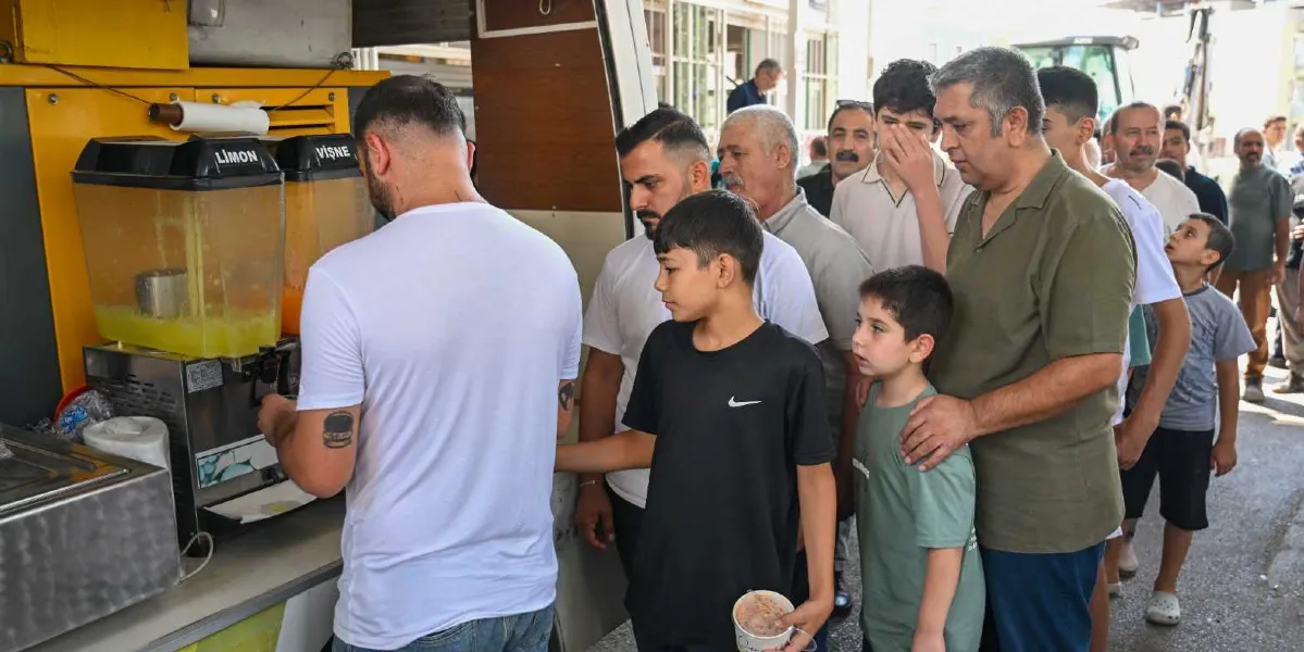 Bornova Belediyesi tarafından kurulan bir ikram aracının önünde, limonata ve vişne suyu almak için sıra bekleyen çocuklar ve yetişkinler. Beyaz tişörtlü bir görevli içecek servisi yaparken, ön sırada elinde dondurma kabı tutan bir çocuk ve yanında onu izleyen diğer vatandaşlar görülüyor.