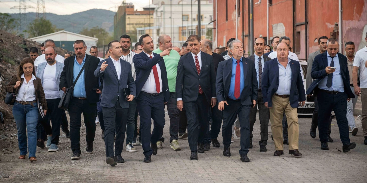 Ağırlıklı olarak takım elbiseli erkeklerden oluşan kalabalık bir grup, açık havada, parke taşlı bir yolda yürüyerek ilerliyor. Grubun ön sıralarında yer alan iki kişi bir yöne doğru işaret ederek bir şeyler anlatıyor. Arka planda toprak bir alan, ağaçlar ve eski bir bina yapısı görülüyor. Grubun genelinde resmi bir kıyafet tarzı hakimken, bazı kişiler daha günlük kıyafetler tercih etmiş.