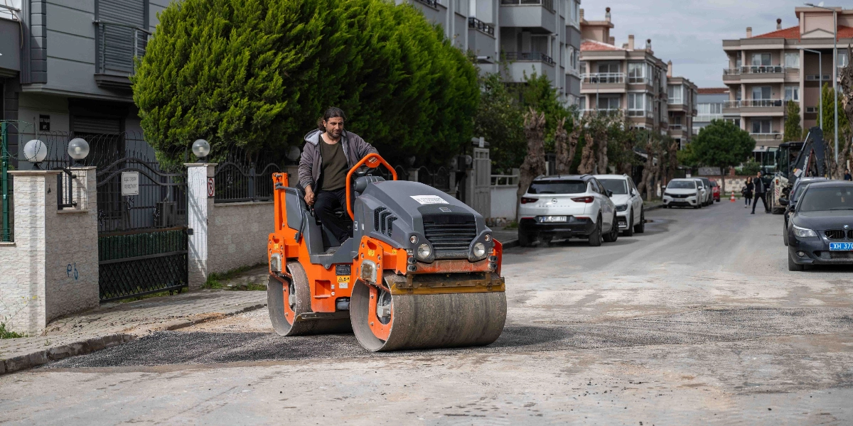 Bornova'da bir yerleşim bölgesindeki sokakta, turuncu renkli bir silindir operatörü asfalt düzeltme çalışması yapıyor. Operatör, gri bir kapüşonlu ceket giymiş şekilde iş makinesini kullanırken, arka planda park halindeki araçlar ve apartmanlar görülüyor.